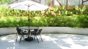 A round table with four chairs and a white umbrella in a sunlit outdoor area with green plants in the background. - Comfy And Classic Studio At Grand Sungkono Lagoon Apartment (Surabaya)
