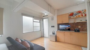 Un espace de vie compact comprenant un canapé, un coin cuisine avec des meubles en bois, une télévision et une fenêtre avec des stores.