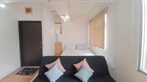 Une pièce de taille modeste comprenant un lit, un canapé sombre agrémenté de coussins, un petit meuble et une fenêtre avec des stores.