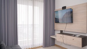 A modern living room with a flat-screen TV mounted on a wooden wall, a grey sofa, and a large window with sheer curtains. - Well Designed 1Br Apartment At M-Town Signature (Gading Serpong)