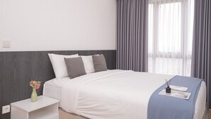 A hotel room with a bed, a nightstand, a vase with flowers, and a window with curtains. - Well Designed 1Br Apartment At M-Town Signature (Gading Serpong)