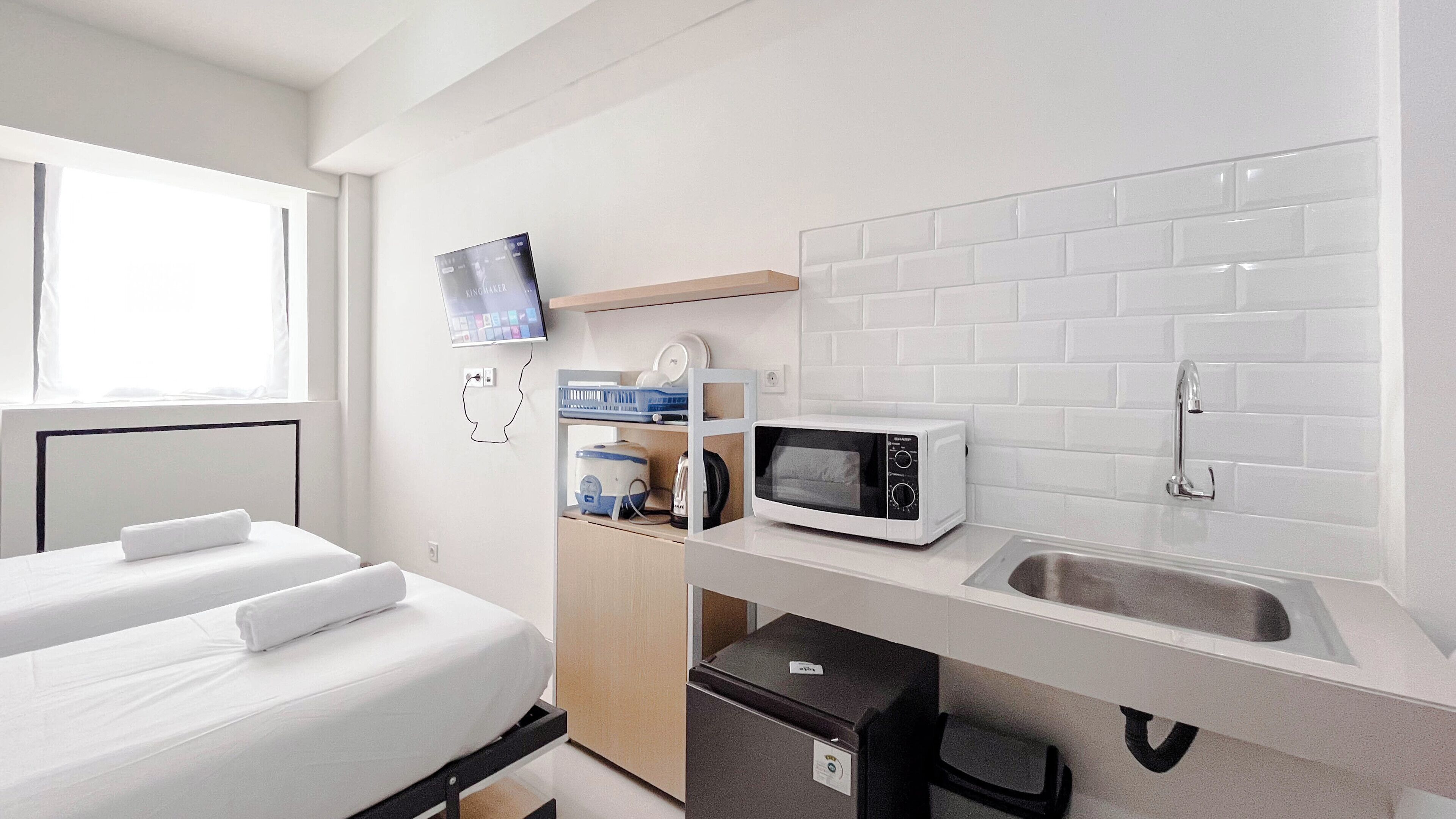 A compact hotel room with a bed, a small kitchenette, a microwave, a sink, and a television mounted on the wall.