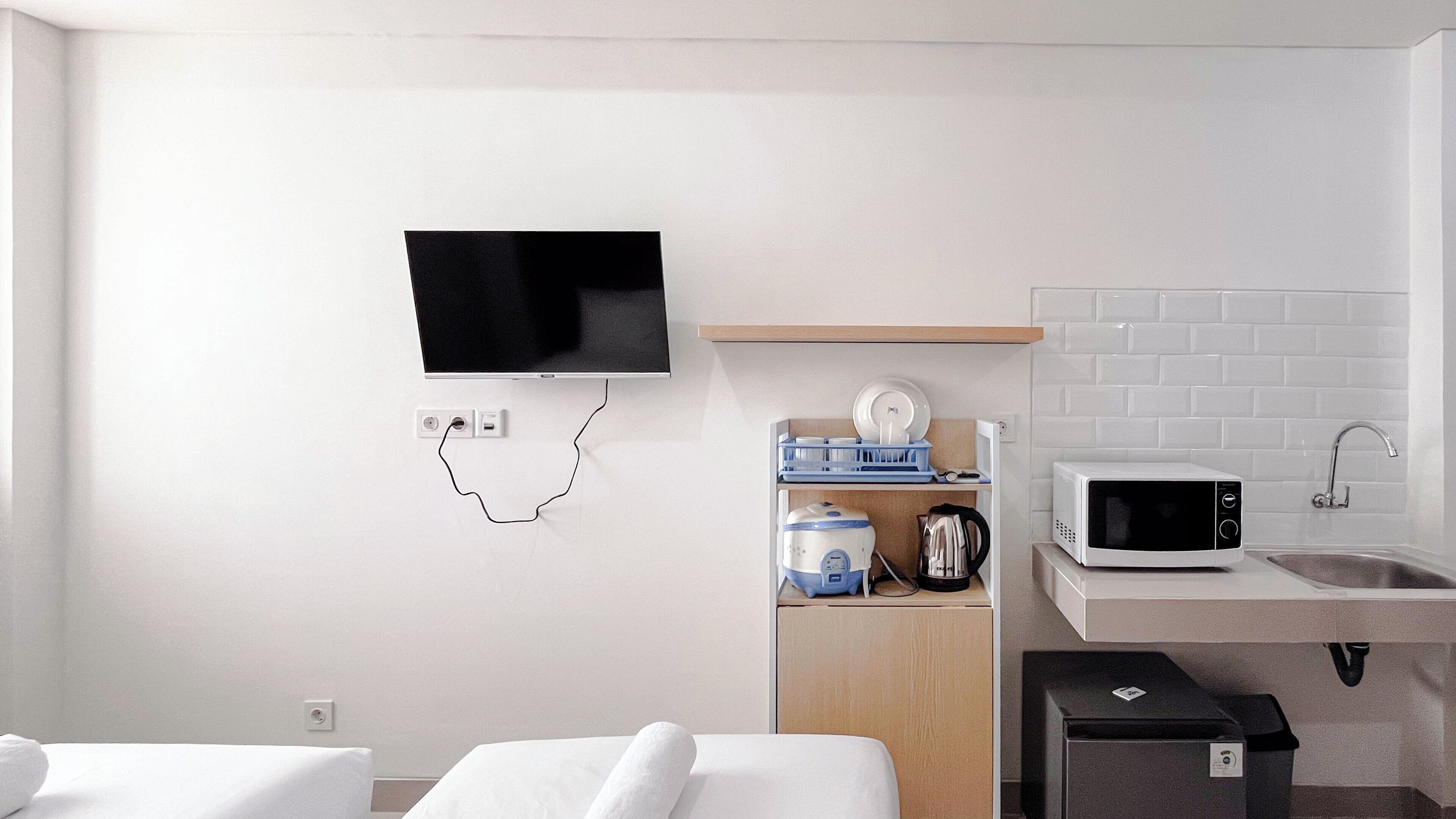 A modern hotel room with a flat-screen TV mounted on the wall, a wooden shelf with kitchen appliances, a microwave, and a sink.