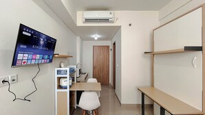 A compact room with a TV mounted on the wall displaying a movie, a small desk with a chair, a microwave, and a cabinet with storage bins. - Comfortable Studio Room Apartment (No Kitchen) At Bandaraya Tallasa City Makassar (Makassar)