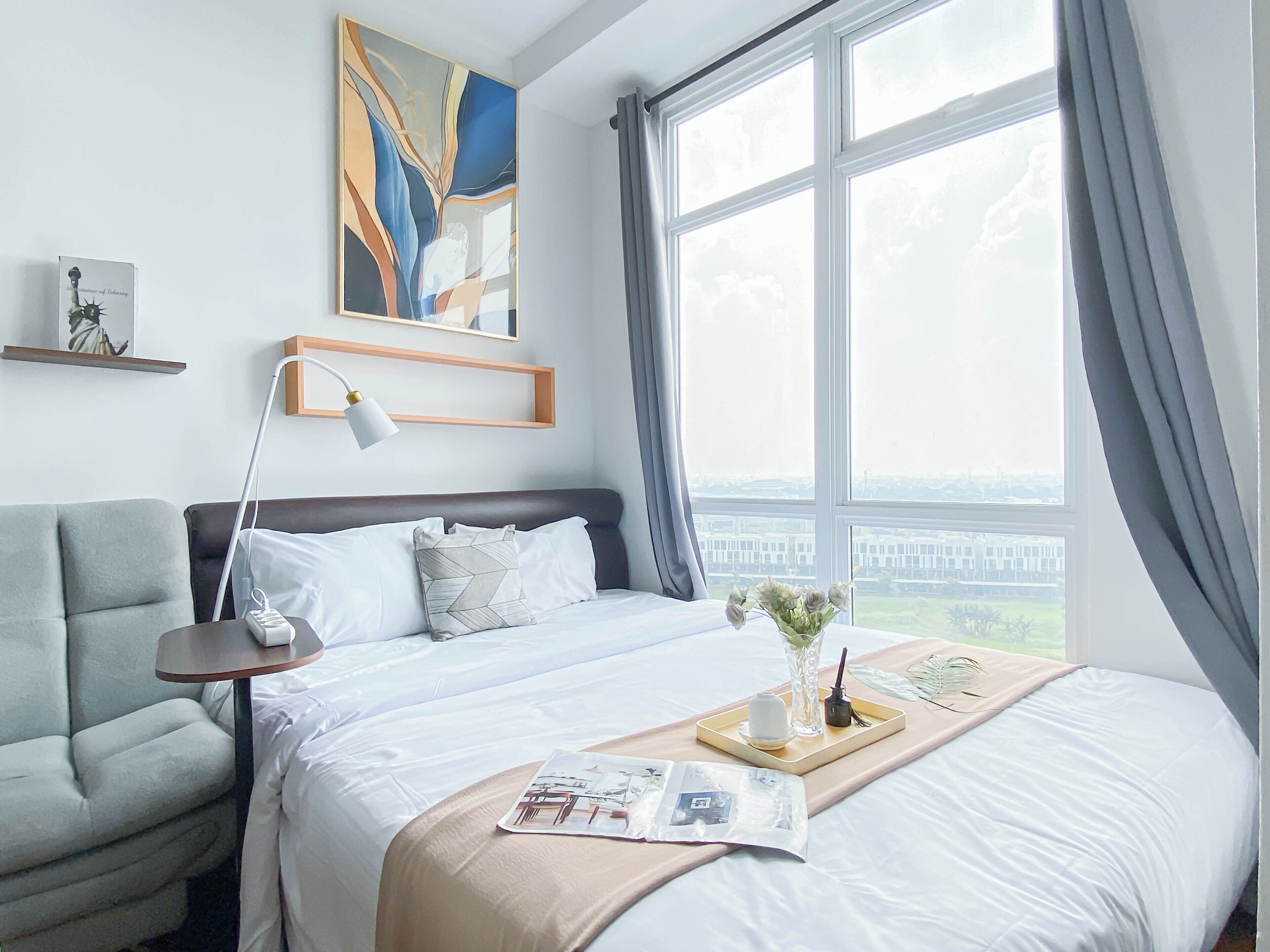 A hotel room with a bed, a desk, a chair, a lamp, a window with curtains, and a painting on the wall.