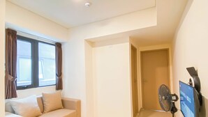 A modern living room with a beige sofa, a wooden side table, a standing fan, and a mounted TV. - Best Choice And Comfy 2Br At Apartment Meikarta (Cikarang)