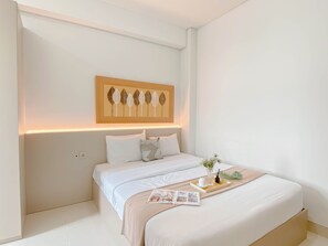 A neatly made bed with a beige bedspread, a decorative wall art piece above it, and a bedside table with a vase of flowers and a magazine. - Homey And Tranquil 2Br Apartment Transpark Cibubur (Depok)
