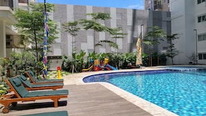 A swimming pool with a wooden deck and lounge chairs, surrounded by greenery and a playground.