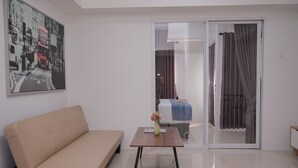 A modern living room with a beige sofa, a small wooden table with a vase of flowers, and a glass door leading to a balcony. - Elegant And Homey 1Br Apartment Tamansari Bintaro Mansion (South Tangerang)