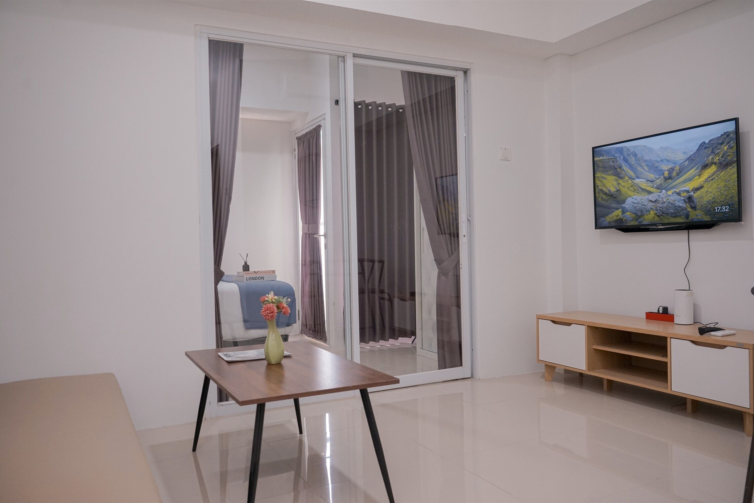 Ein modernes Wohnzimmer mit einer Schiebeglastür, einem flachbildfernseher an der Wand, einem Holztisch mit einer Vase und einem weißen Schrank mit einem roten Gegenstand darauf.