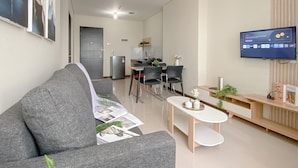 A modern living room with a grey sofa, a dining area with a small table and chairs, a kitchen in the background, and a TV mounted on the wall. - Minimalistic Designed 2Br Nifarro Park Apartment (Jakarta)