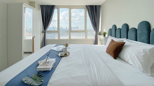 A hotel room with a large bed, a view of the city through the window, and a bedside table with a lamp and a plant. - Minimalistic Designed 2Br Nifarro Park Apartment (Jakarta)