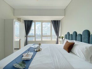 Ein Hotelzimmer mit einem großen Bett, Blick auf die Stadt durch das Fenster und einem Nachttisch mit Lampe und Pflanze.