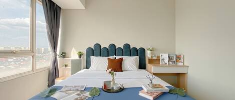 A neatly made bed with a blue bedspread, white pillows, and a decorative pillow. There is a tray with a vase and a book on the bed. A window with curtains is visible, and a nightstand with a lamp and a small plant is next to the bed.