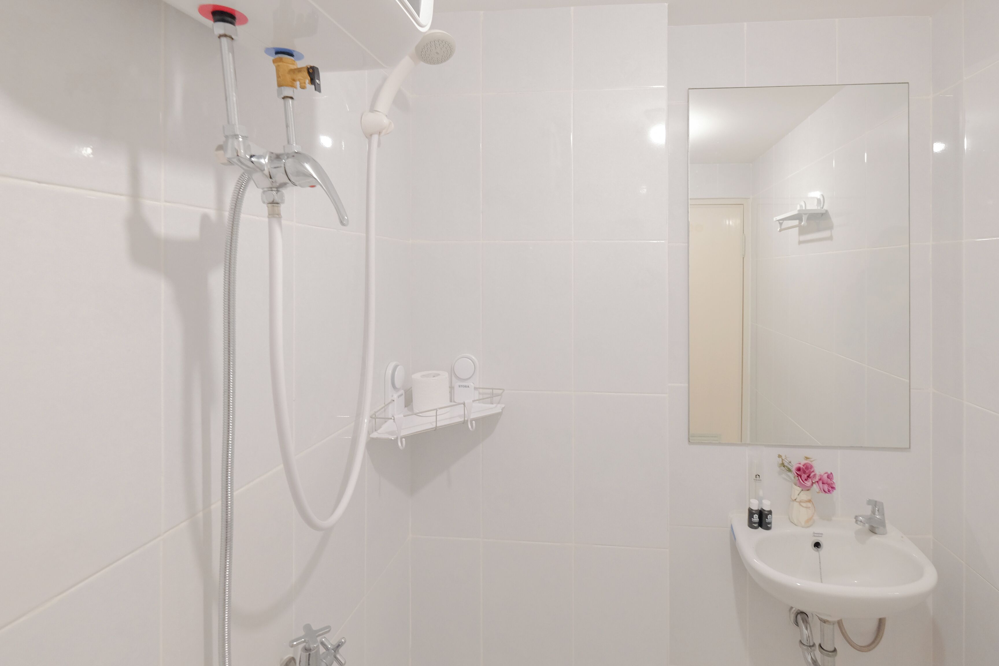 A bathroom with white tiles, a shower with a handheld showerhead, a sink with a mirror above it, and toiletries on the sink.