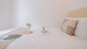 A neatly made bed with a beige headboard, a small bedside table with a vase of flowers, and a book. - Spacious And Homey Corner 2Br Tokyo Riverside Pik 2 Apartment (Teluknaga)