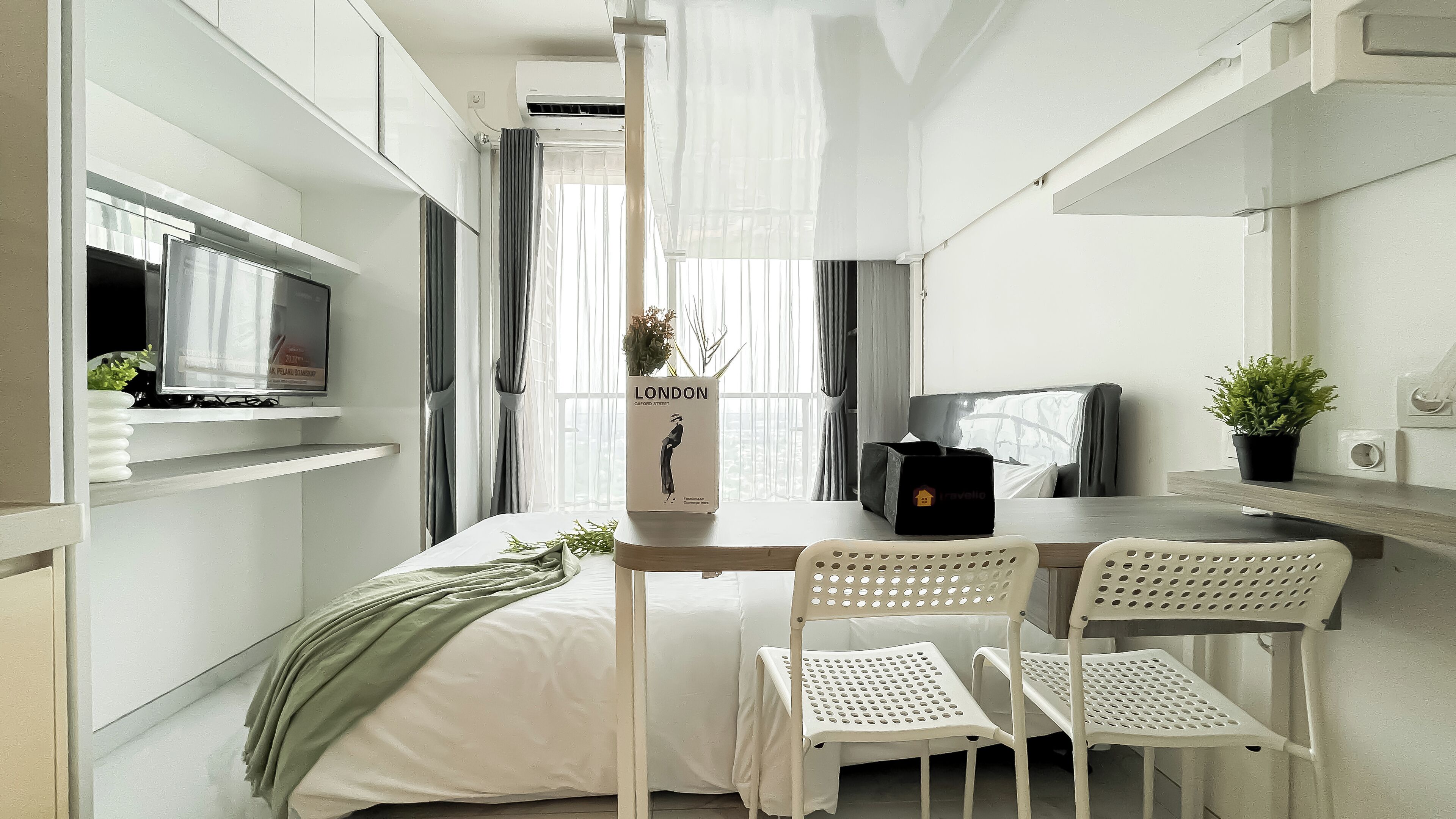 A compact, modern room with a bed, a small table, two chairs, a TV, and a potted plant.