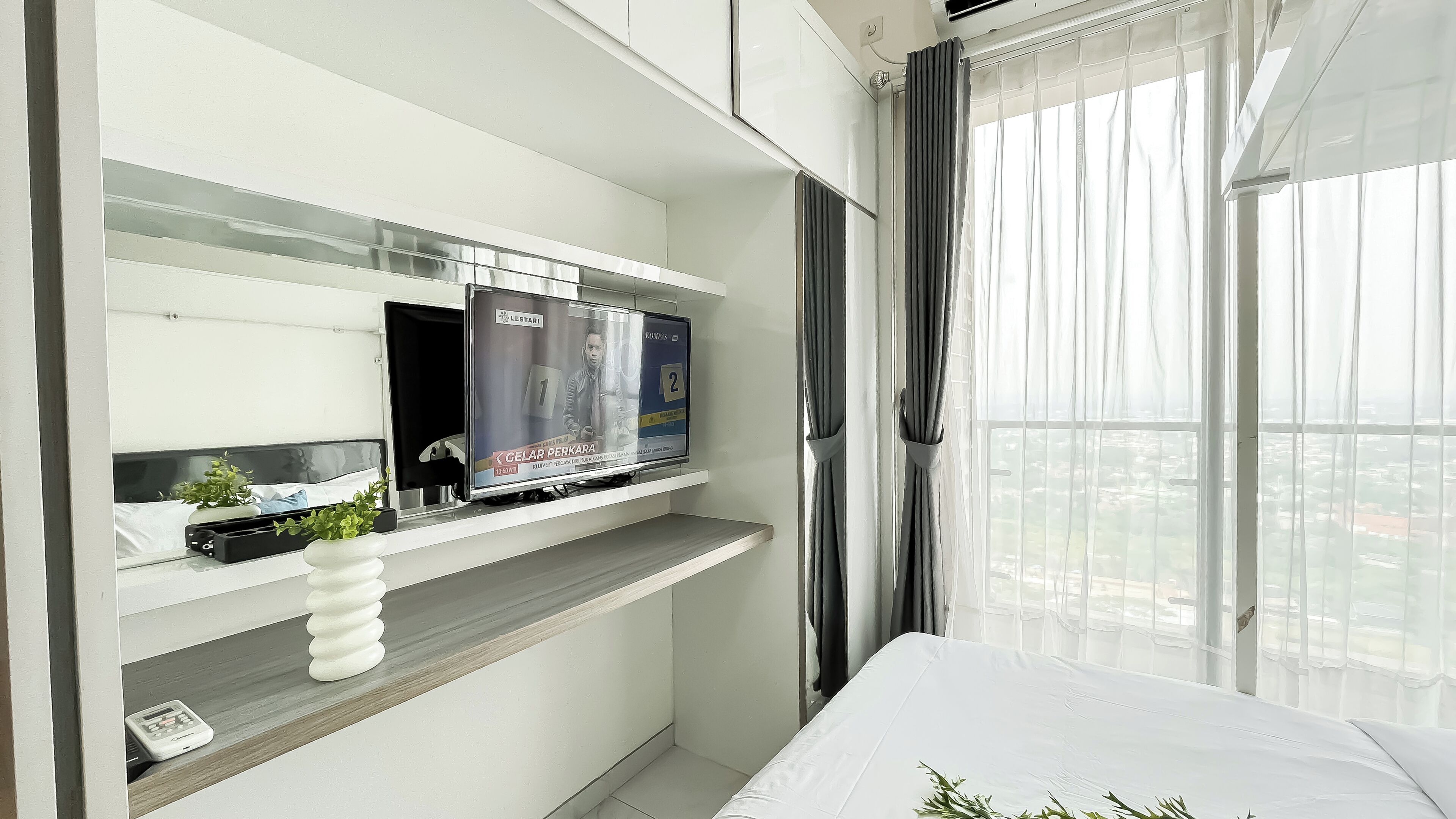 A modern hotel room with a TV on a shelf, a small plant, and a view of the city through large windows.