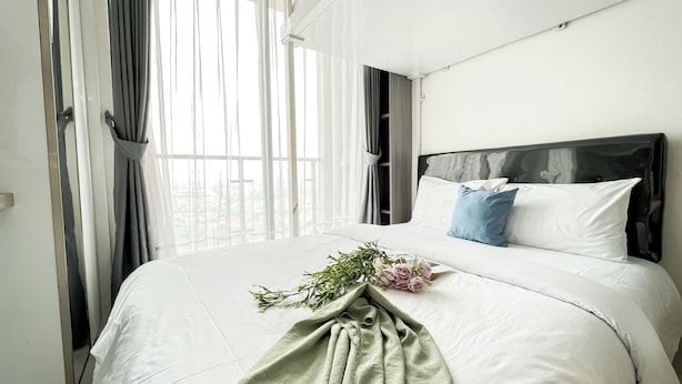 A hotel room with a large bed, white bedding, a headboard, and sheer curtains. - Modern Look And Homey Studio At Apartment Sky House Bsd (Pagedangan)