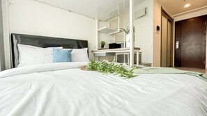 A neatly made bed with white bedding and a blue pillow, a desk with a computer and a small plant, and a dark door in the background.
