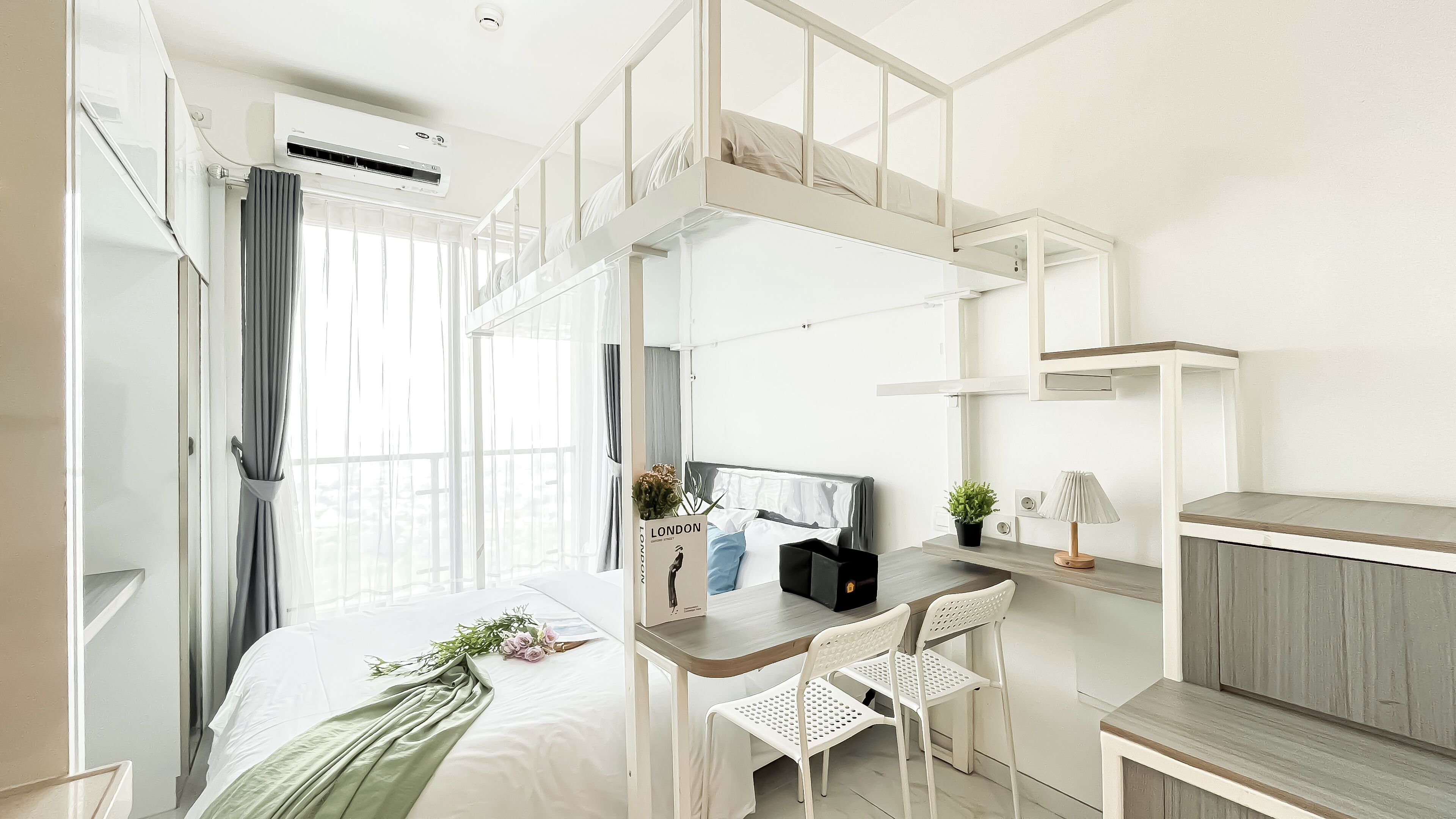 A modern hotel room with a bunk bed, a desk with a chair, a small table, a potted plant, and a lamp.