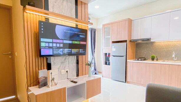 A modern living room with a flat-screen TV mounted on a wooden wall, a built-in kitchen area with a refrigerator and a sink, and a grey sofa. - Best Living 2Br At Meikarta Apartment (Cikarang)
