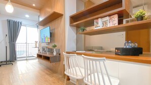 A modern living room with a wooden floor, a TV mounted on the wall, a balcony with a view, and a dining area with white chairs. - Great Choice 1Br Apartment At Green Bay Condominium (Jakarta)