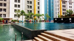 A swimming pool area with steps leading into the water, surrounded by palm trees and a multi-story building with balconies. - Comfy And Modern 1Br Apartment Paramount Skyline (Gading Serpong)