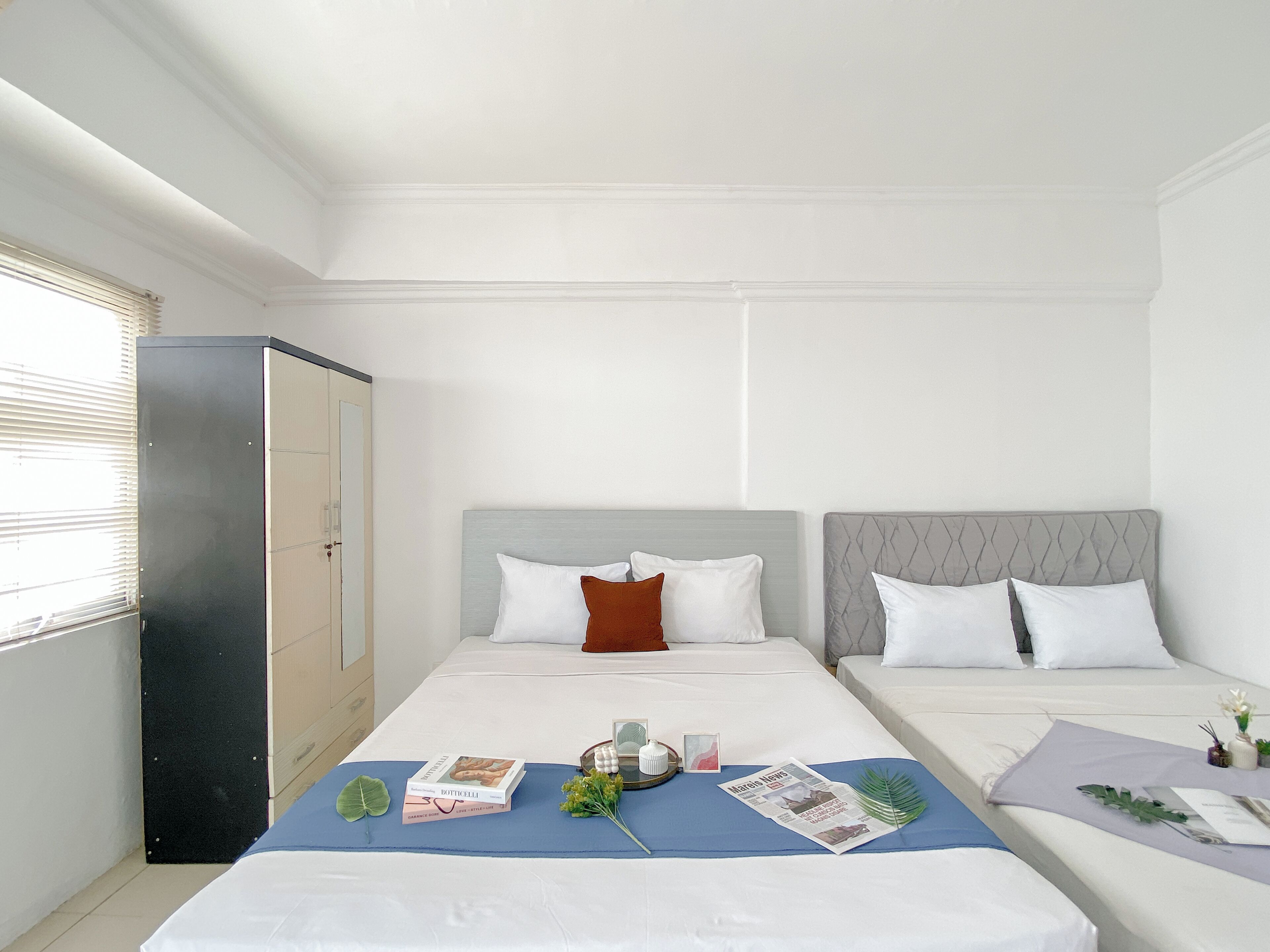 Une chambre d’hôtel avec deux lits, une armoire et une table de chevet sur laquelle se trouvent un livre, des fleurs et un journal.