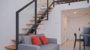 A modern living room with a grey sofa, red cushions, a wooden staircase, and a dining area with a table and chairs. - Spacious And Homey Studio Loft At Kingland Avenue Apartment (South Tangerang)