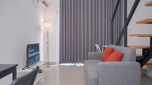 A modern living room with a grey sofa, a TV on a stand, a lamp, and a staircase. - Spacious And Homey Studio Loft At Kingland Avenue Apartment (South Tangerang)