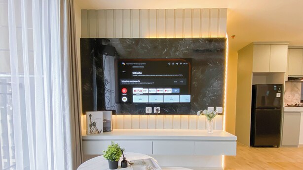 A modern kitchen with a built-in TV, a dining area with a round table, and a refrigerator. - Modern Look And Restful 3Br At Meikarta Apartment (Cikarang)