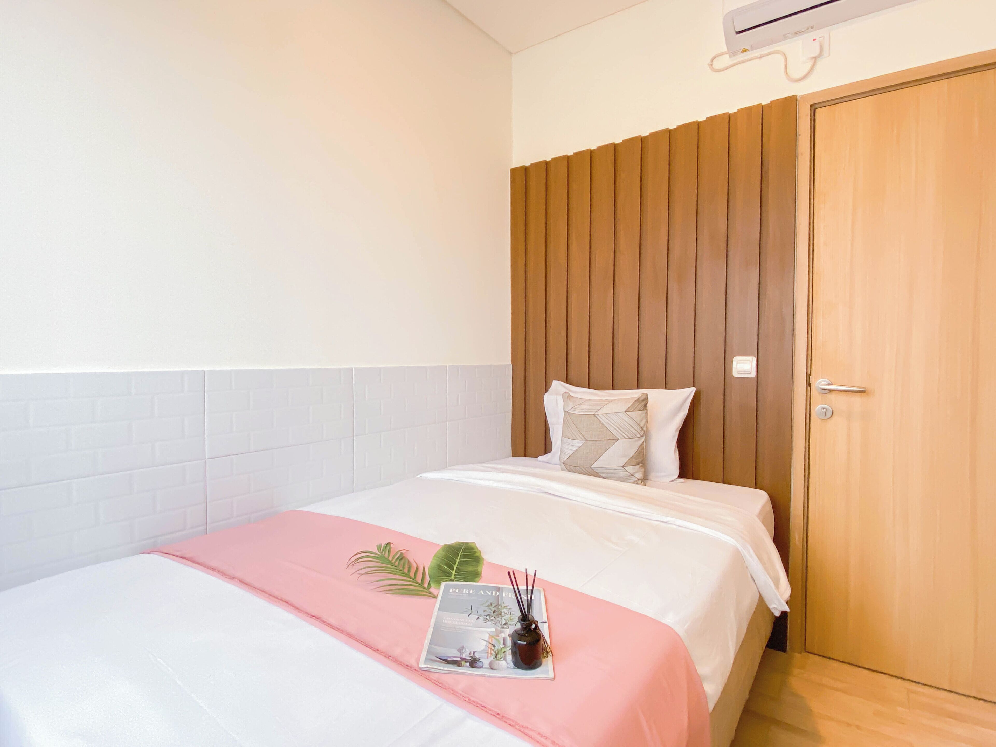 Habitación de hotel con cabecera de madera, cama con ropa blanca y un toque de color rosa, una planta pequeña y una revista sobre la cama.