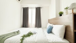 A neatly made bed with white linens, a wooden headboard, and a small bedside table with a lamp and a potted plant. - Warm And Comfy 2Br At Bassura City Apartment (Jakarta)