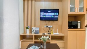 A modern living room with a flat-screen TV mounted on the wall, a wooden cabinet with glass doors, a small table with a magazine and a vase of flowers, and a decorative plant. - Cozy And Minimalist 1Br With Working Room Meikarta Apartment (Cikarang)