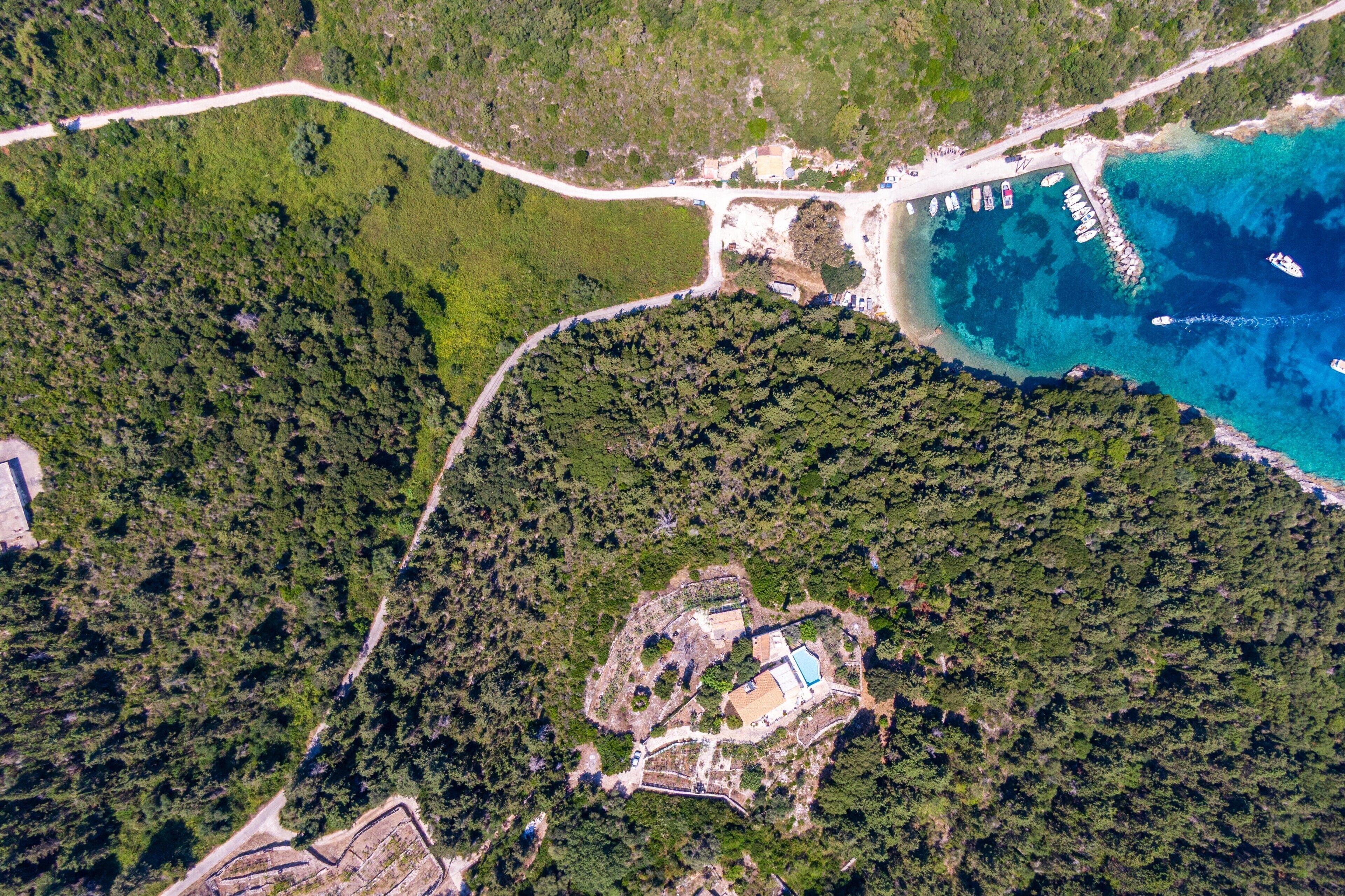 Une vue aérienne d’une zone côtière avec une forêt dense, une route dégagée, un quai avec des bateaux et un bâtiment doté d’une piscine.