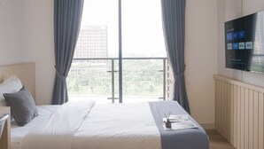 A hotel room with a bed, a desk, a flat-screen TV mounted on the wall, and a view of a cityscape through large windows. - Best Deal Studio Room Apartment At Sky House Bsd (Pagedangan)