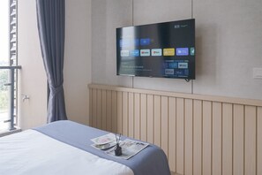 A hotel room with a bed, a flat-screen TV mounted on the wall displaying various streaming services, a window with curtains, and a wooden headboard. - Best Deal Studio Room Apartment At Sky House Bsd (Pagedangan)