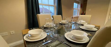 A dining table set with white plates, bowls, and cutlery, surrounded by beige chairs, with windows and curtains in the background.