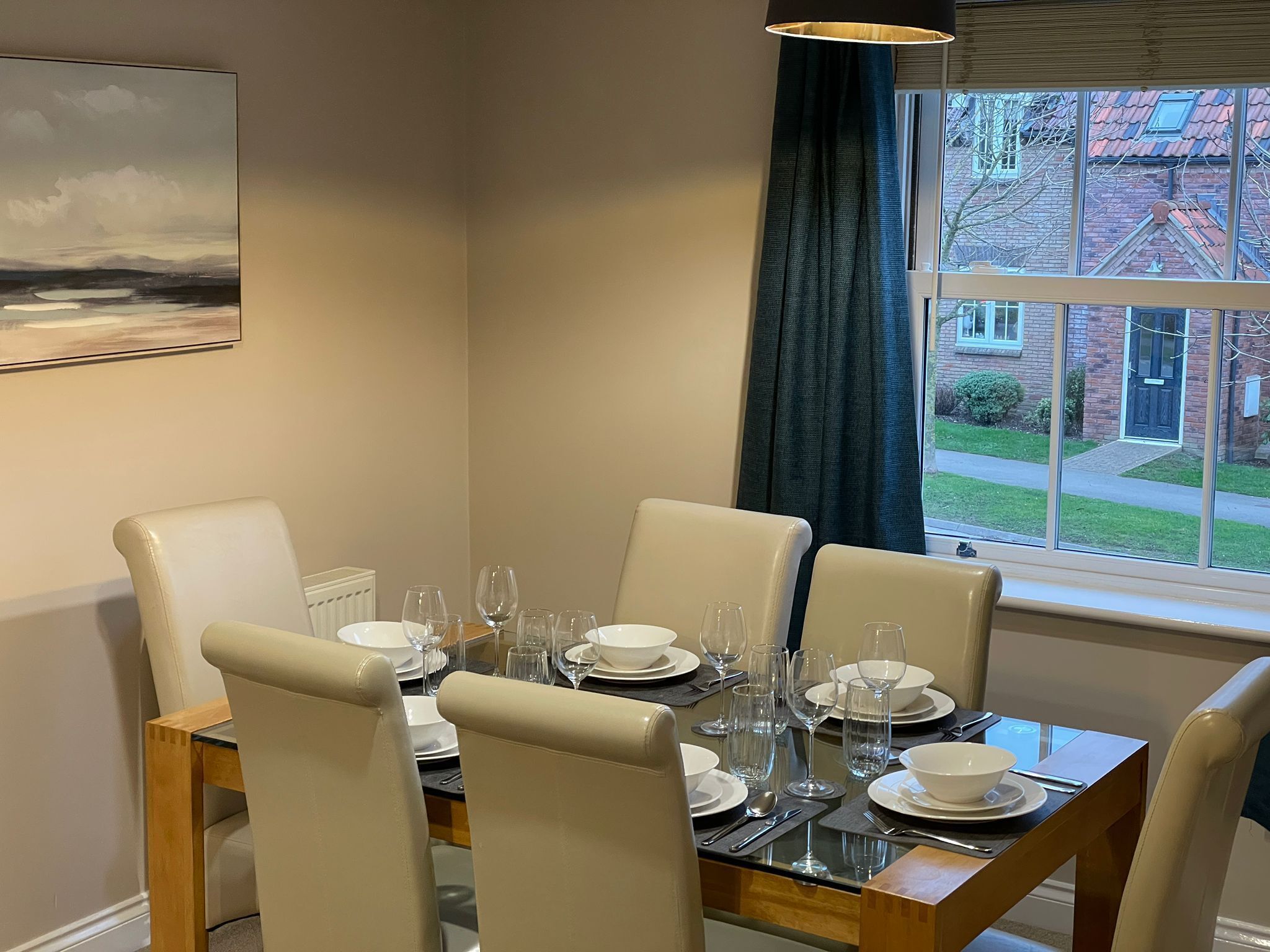 A dining table set for four with chairs, a window with curtains, and a painting on the wall.