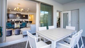 Une cuisine moderne avec des meubles de couleur sombre et un plan de travail blanc, un coin repas avec une table et des chaises blanches, et une porte-fenêtre coulissante donnant sur une terrasse extérieure.