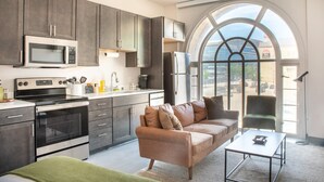 Et moderne køkken med mørke skabe, en mikrobølgeovn, komfur og køleskab. Et stort, buet vindue giver udsigt over byen. Der er en beige sofa og et lille bord i opholdsstuen.