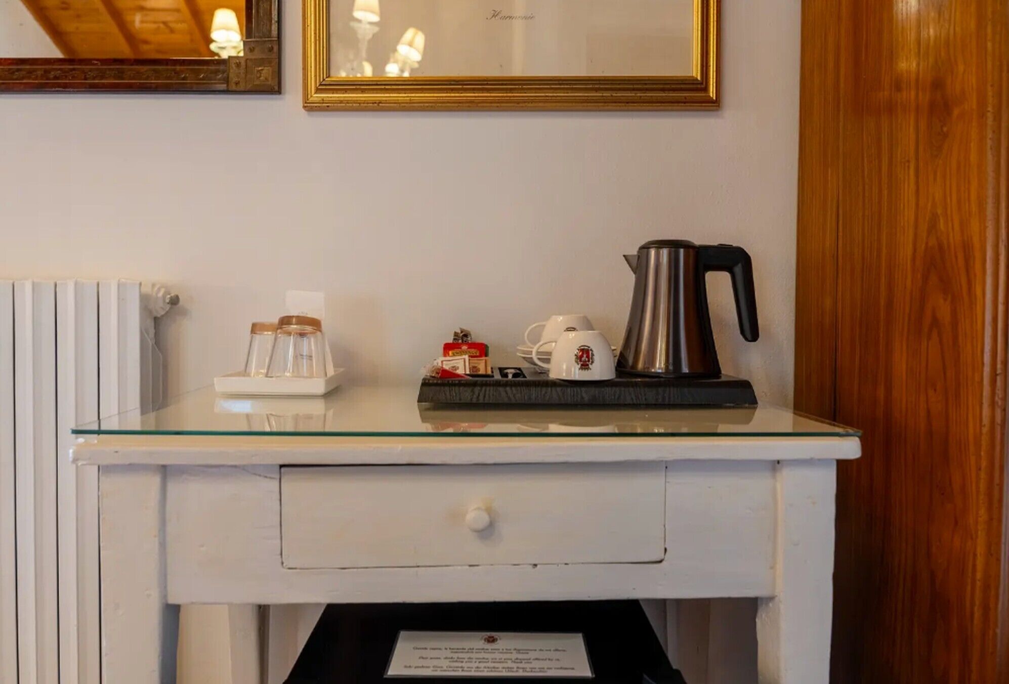 Uma secretária branca com bancada de vidro, uma máquina de café, uma chaleira e um pequeno tabuleiro com um bule e uma caixa de saquinhos de chá.