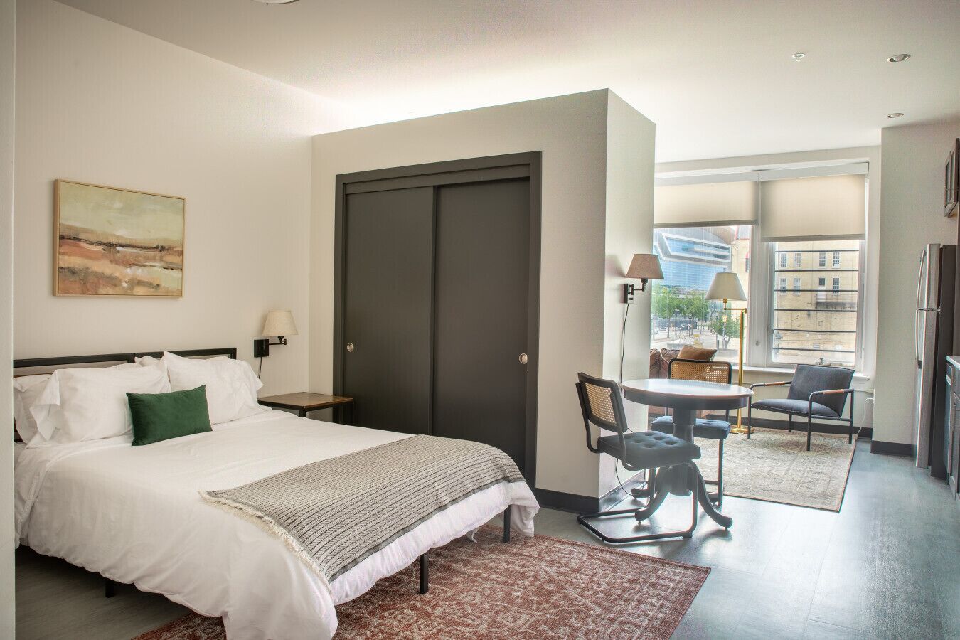 A hotel room with a bed, a desk, a chair, a table, and a view of the city through large windows.
