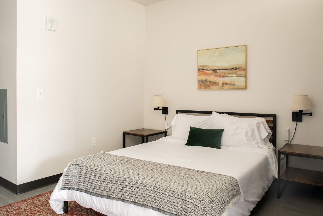 Un dormitorio con una cama, dos mesitas de noche, una lámpara de pared, un cuadro y una alfombra estampada.