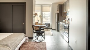 A modern hotel room with a bed, a dining area with a table and chairs, a kitchen with cabinets and appliances, and a large window providing natural light. - Deer District Studio Apt - One Block from Fiserv and Baird (Milwaukee)