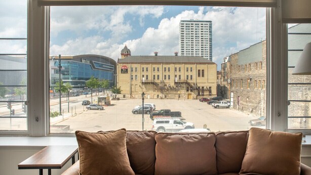 A room with a large window overlooking a city street, a brown leather sofa, and a small wooden table. - Deer District Studio Apt - One Block from Fiserv and Baird (Milwaukee)