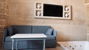 A room with a stone arch ceiling, a grey sofa, a white table, and a white bathtub. - King Room with Jacuzzi - Charming in Felline - Borgo Don Ciccio (Felline)