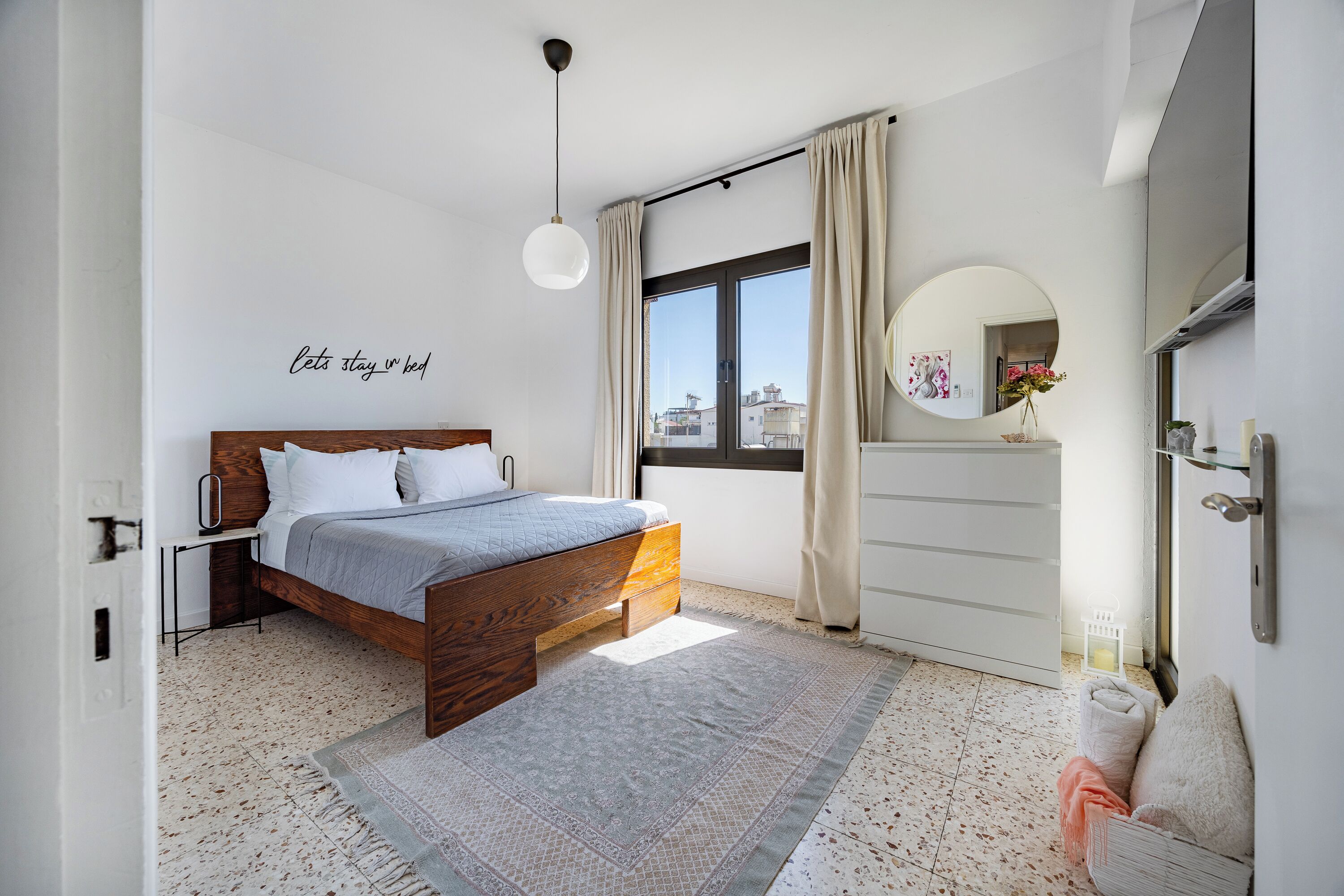 A bedroom with a bed, a bedside table, a dresser, a mirror, and a window with curtains.