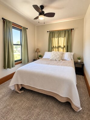 Un dormitorio con cama, ventilador de techo, ventana con cortinas, mesita de noche y lámpara.